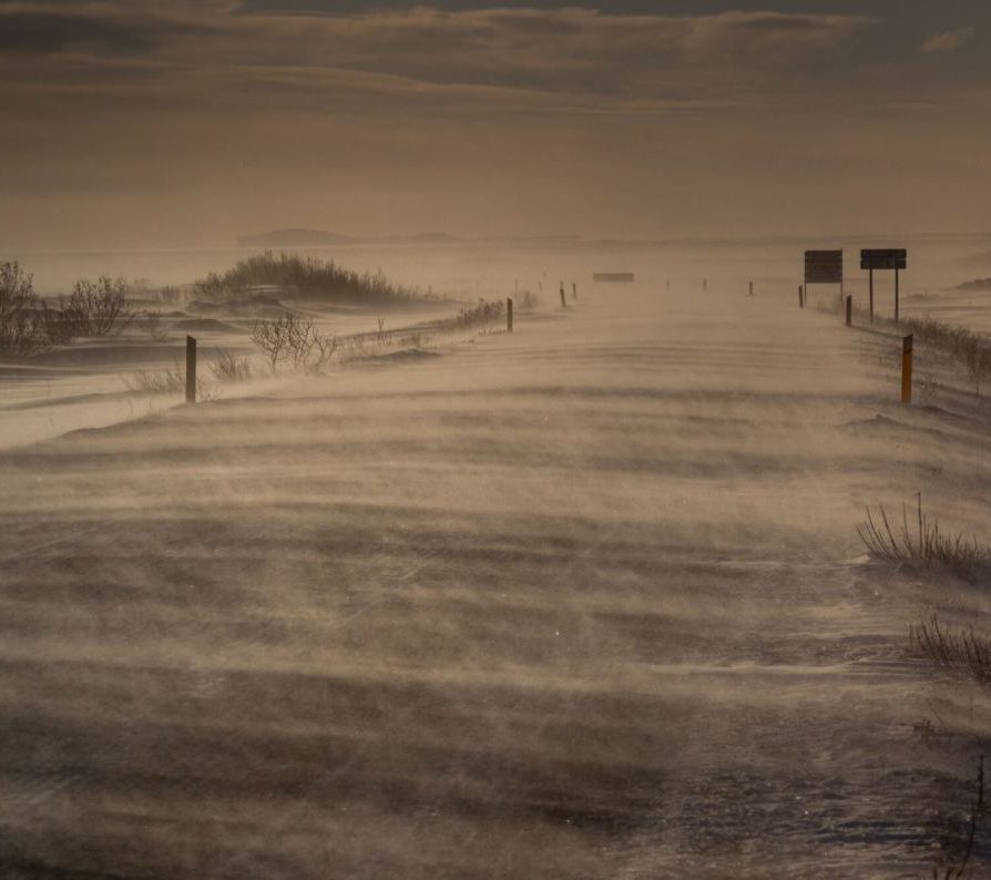 Myndin sýnir frá Suðurlandi í slæmu skyggni þar sem óveður dregur yfir landið. Mikið snjófok er við veginn og erfitt er að lesa á skilti. Sést glitta í Vestmannaeyjar örlítið í bakgrunninum.
