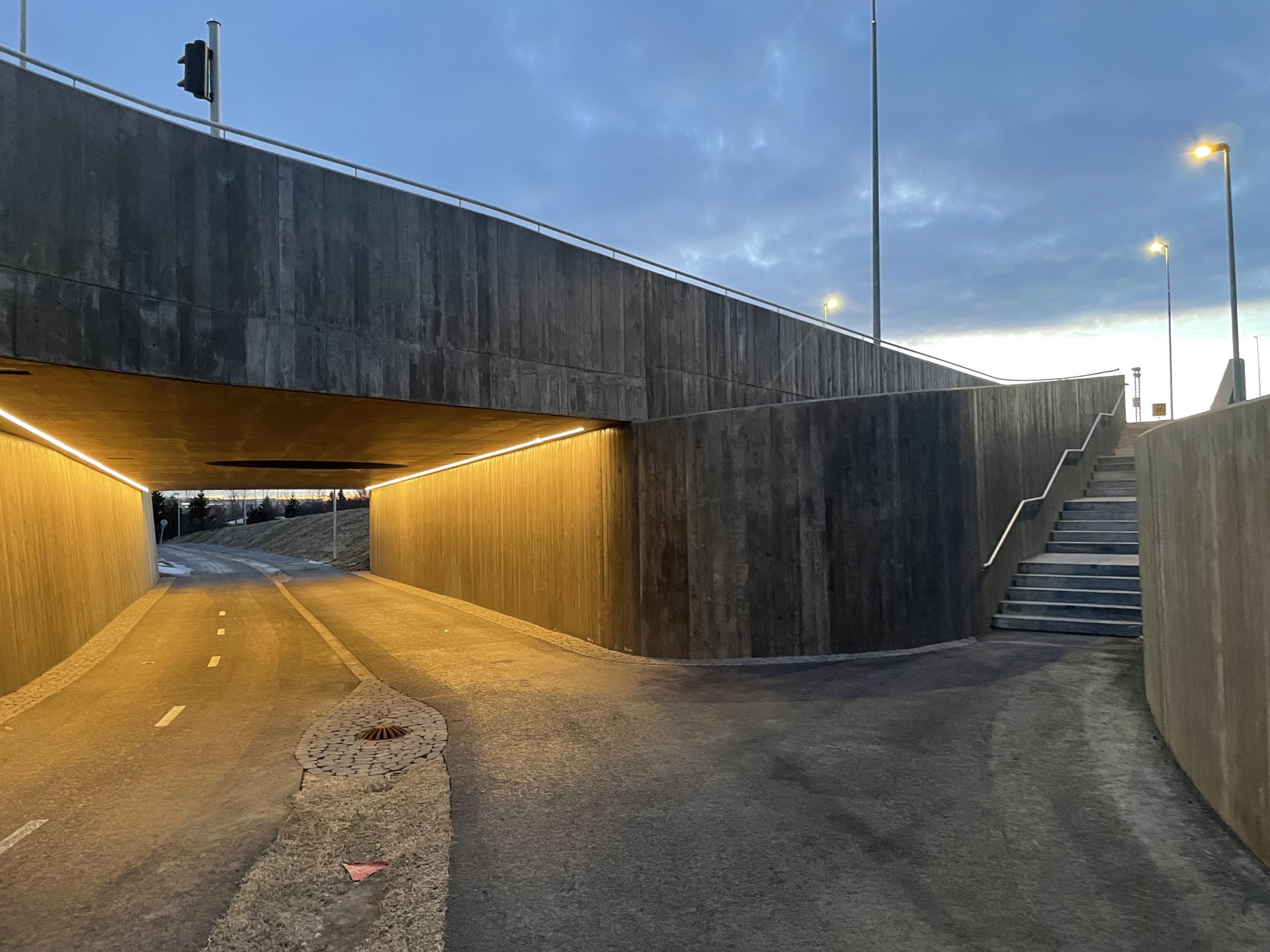 Myndin sýnir undirgöng við Arnarnes. Göngin eru í nútíma- brútalismahönnun. Göngin eru lýst upp í mildum tónum, og gat til þess að hleypa inn sólarljósi er staðsett í miðju gangnaþaki.