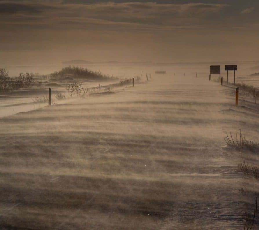 Myndin sýnir frá Suðurlandi í slæmu skyggni þar sem óveður dregur yfir landið. Mikið snjófok er við veginn og erfitt er að lesa á skilti. Sést glitta í Vestmannaeyjar örlítið í bakgrunninum.